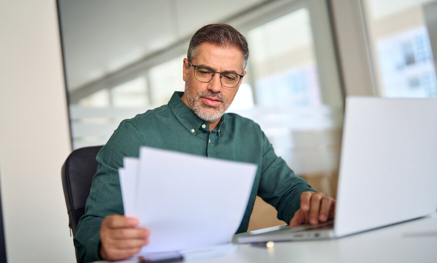 Ausbilder checkt Papiere am Schreibtisch mit Laptop