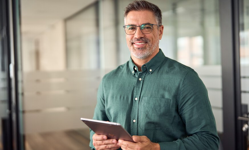 Mann in mittlerem Alter im lockeren Hemd hält Tablet in der Hand und lächelt in die Kamera