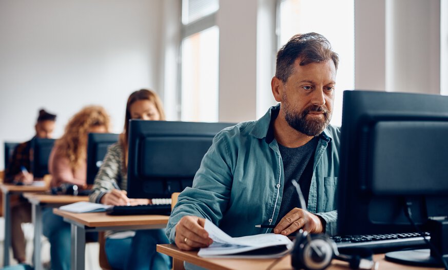 Mann mittleren Alters im blauen Jeanshemd sitz hinter Computer und lernt